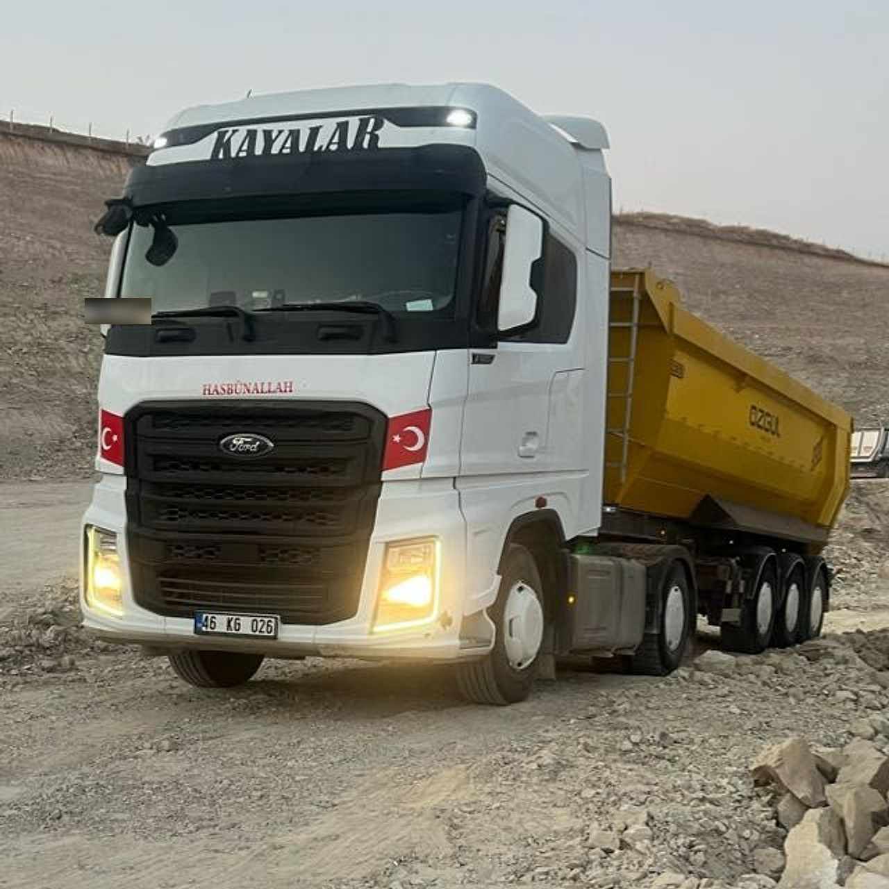 Ön camının üstünde büyük harflerle KAYALAR yazan beyaz Ford çekici ve sarı damperli dorse, farları yanık